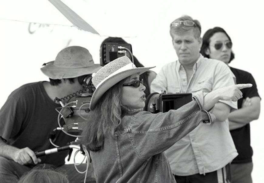 A black and white image of film director Susan Seidelman sitting in front of a camera with a group of 3 people. She is pointing to the right